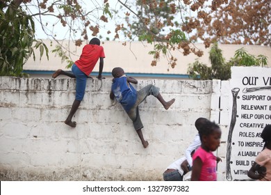 Sukuta, The Gambia, Africa, May 18, 2017:   Horizontal Photo Of A Group African Children In Colorful  Clothes, Climbing A White Wall With Written School  Mission Statement,  Outdoors On A Sunny Day 