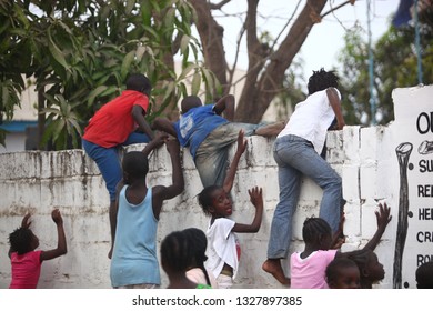 Sukuta, The Gambia, Africa, May 18, 2017:   Horizontal Photo Of A Group African Children In Colorful  Clothes, Climbing A White Wall With Written School  Mission Statement,  Outdoors On A Sunny Day 