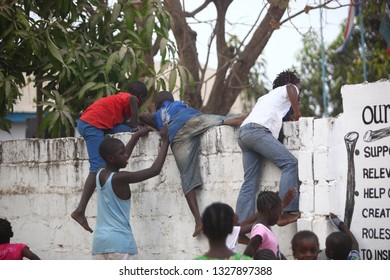 Sukuta, The Gambia, Africa, May 18, 2017:   Horizontal Photo Of A Group African Children In Colorful  Clothes, Climbing A White Wall With Written School  Mission Statement,  Outdoors On A Sunny Day 