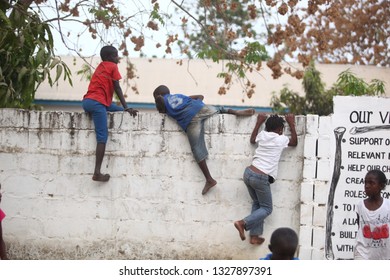 Sukuta, The Gambia, Africa, May 18, 2017:   Horizontal Photo Of A Group African Children In Colorful  Clothes, Climbing A White Wall With Written School  Mission Statement,  Outdoors On A Sunny Day 