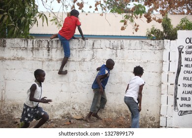 Sukuta, The Gambia, Africa, May 18, 2017:   Horizontal Photo Of A Group African Children In Colorful  Clothes, Climbing A White Wall With Written School  Mission Statement,  Outdoors On A Sunny Day 