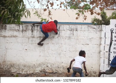 Sukuta, The Gambia, Africa, May 18, 2017:   Horizontal Photo Of A Group African Children In Colorful  Clothes, Climbing A White Wall With Written School  Mission Statement,  Outdoors On A Sunny Day 