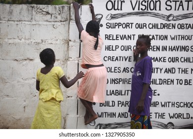 Sukuta, The Gambia, Africa, May 18, 2017: Horizontal Photo Of A Girl In Pink Clothes Climbing A White Wall With Written School  Mission Statement,  Outdoors On A Sunny Day, With Other Children Around