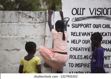 Sukuta, The Gambia, Africa, May 18, 2017: Horizontal Photo Of A Girl In Pink Clothes Climbing A White Wall With Written School  Mission Statement,  Outdoors On A Sunny Day, With Other Children Around