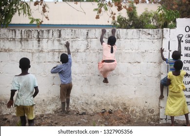 Sukuta, The Gambia, Africa, May 18, 2017: Horizontal Photo Of A Girl In Pink Clothes Climbing A White Wall With Written School  Mission Statement,  Outdoors On A Sunny Day, With Other Children Around