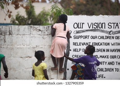 Sukuta, The Gambia, Africa, May 18, 2017: Horizontal Photo Of A Girl In Pink Clothes Climbing A White Wall With Written School  Mission Statement,  Outdoors On A Sunny Day, With Other Children Around