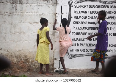 Sukuta, The Gambia, Africa, May 18, 2017: Horizontal Photo Of A Girl In Pink Clothes Climbing A White Wall With Written School  Mission Statement,  Outdoors On A Sunny Day, With Other Children Around