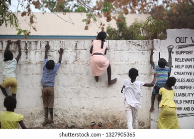 Sukuta, The Gambia, Africa, May 18, 2017: Horizontal Photo Of A Girl In Pink Clothes Climbing A White Wall With Written School  Mission Statement,  Outdoors On A Sunny Day, With Other Children Around