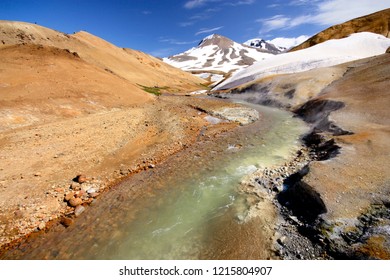 Sulfuric Landscape In Iceland With Green - Yellow River Flowing In A Orange Sand And Stones And Snow And Mountain In The Background