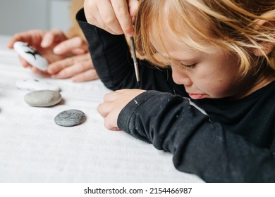 Sulking Little Boy Painting On His Hand With A Brush. Close Up, Side View. Hands Of His Mother In Background. She Is Holding Stone With A Painted Shape.