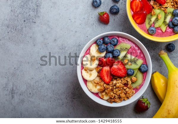 Summer acai smoothie bowls with strawberries,\
banana, blueberries, kiwi fruit and granola on gray concrete\
background. Breakfast bowl with fruit and cereal, close-up, top\
view, space for text