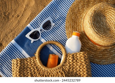 Summer accessories on sandy beach at sunset. Woven bag with sunscreen inside next to stylish sun hat. White sunglasses lie on blue striped towel. Vacation essentials for sun protection. - Powered by Shutterstock