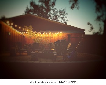 Summer Backyard At Night With Adirondack Chairs Surrounding A Fire Pit With Edison Light Bulbs Glowing Against Dark Trees And A Sunset Sky With A Flat Vintage Look