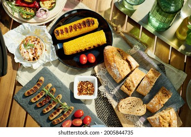 Summer Backyard Picnic. Upper View Of Desk With Electric Grill, Grilled Sausages, Corn, Bread And Camembert.