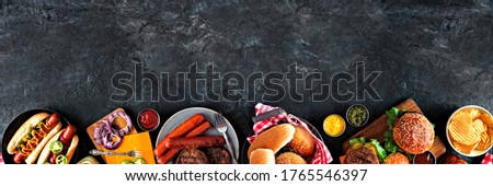 Summer BBQ food table scene with hot dog and hamburger buffet. Overhead view bottom border over a dark slate background. Copy space.