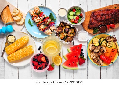 Summer BBQ Or Picnic Food Concept. Assortment Of Grilled Meats, Vegetables, Fruits, Salad And Potatoes. Top Down View Table Scene With A White Wood Background.