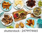 Summer BBQ or picnic food table scene. Burgers, grilled meats, potatoes, smoked corn, fruits, salad and snacks. Overhead view on a white wood background.