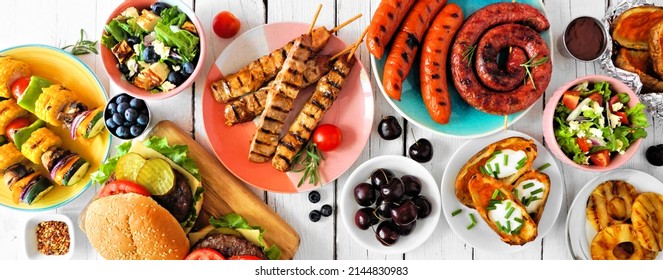 Summer BBQ or picnic food table scene. Selection of burgers, grilled meat, vegetables, fruits, salad and potatoes. Top down view on a white wood banner background.