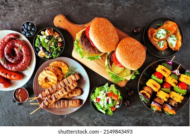 Summer BBQ Or Picnic Food Table Scene. Mixture Of Burgers, Grilled Meat, Vegetables, Fruits, Salad And Potatoes. Overhead View On A Dark Background.