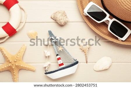 Summer beach flat lay featuring straw sun hat, black sunglasses, seashells, starfish, decorative sailboat and lighthouse accent on light wooden background. Top view