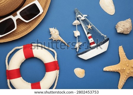 Summer beach flat lay featuring straw sun hat, black sunglasses, seashells, starfish, decorative sailboat and lighthouse accent on dark blue background. Top view