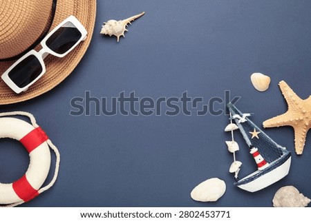Summer beach flat lay featuring straw sun hat, black sunglasses, seashells, starfish, decorative sailboat and lighthouse accent on dark blue background. Space for text