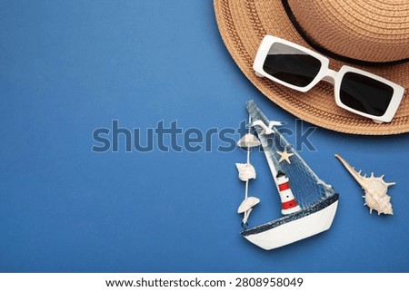 Summer beach flat lay featuring straw sun hat, black sunglasses, seashells, starfish, decorative sailboat and lighthouse accent on dark blue background. Space for text