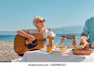 Summer beach picnic. Middle aged couple having picnic at the beach. Bearded man preparing fresh fruit and appetizers for picnic outdoors at seaside, while blond woman playing guitar. - Powered by Shutterstock