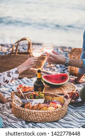 Summer beach picnic at sunset. Young couple having weekend picnic outdoors at seaside with fresh fruit and tray of tasty appetizers, clinking glasses with sparkling wine