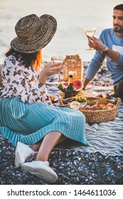 Summer beach picnic at sunset. Young happy couple having weekend picnic outdoors at seaside with fresh fruit and tray of tasty appetizers, drinking sparkling wine and enjoying chat