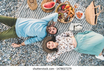 Summer Beach Picnic At Sunset. Young Couple Lying On Striped Blanket, Looking Above At Weekend Picnic Outdoor At Seaside With Sparkling Wine, Fresh Fruit And Tray Of Tasty Appetizers, Top View
