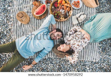 Summer beach picnic at sunset. Young couple lying on striped blanket, looking at each other at weekend picnic outdoor at seaside with sparkling wine, fresh fruit and tray of tasty appetizers, top view