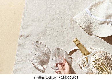 Summer Beach Rest Concept Background With Woman Sun Hat, Crystal Wine Glasses In Hand, Bottle Sparkling Wine In Mesh Bag On Natural Beach Towel As Background, Sunlight Shadows, Pastel Color Lifestyle