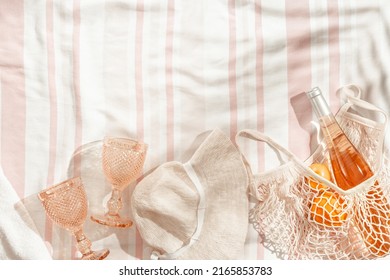 Summer Beach Rest Concept Background With Sun Hat, Color Wine Glasses, Bottle Rose Wine, Orange Fruits In Mesh Bag On Stripes Beach Towel As Background, Sunlight And Palm Leaf Shadows, Pastel Color