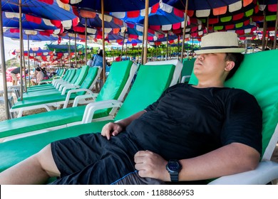 Summer Beach Vacation, Side View Of Asian Man Ware A Hat Relaxing On A Beach Chair Under An Umbrella