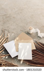 Summer boho wedding stationery still life. Blank greeting cards, envelope mock-ups and silk ribbon. Dry palm leaves on grunge concrete background. Flat lay, top view. Tropical vacation concept. 