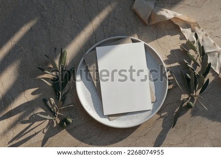 Summer branding, wedding stationery. Blank greeting card, invitation mock up on plate. Beige marble background. Olive tree branches, fruit with silk ribbon in sunlight, long shadows. Flat lay, top.