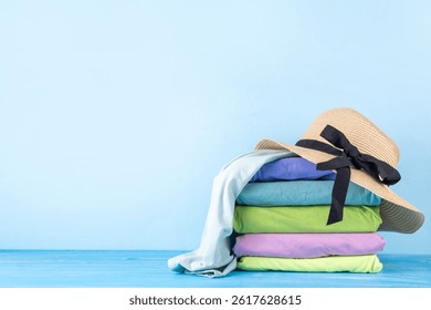 Summer bright clothes pile stack on wooden light blue table, with summer hat, suitcase, shells. Seaside holidays, vacation concept, preparation for travel background