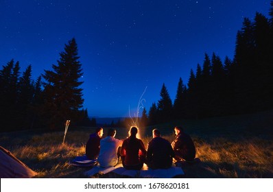 Summer camping under stars. Rear view of group of five hikers, men and woman sitting near bright bonfire, tourist tent under dark night sky with sparkling stars. Concept of tourism, night camping. - Powered by Shutterstock