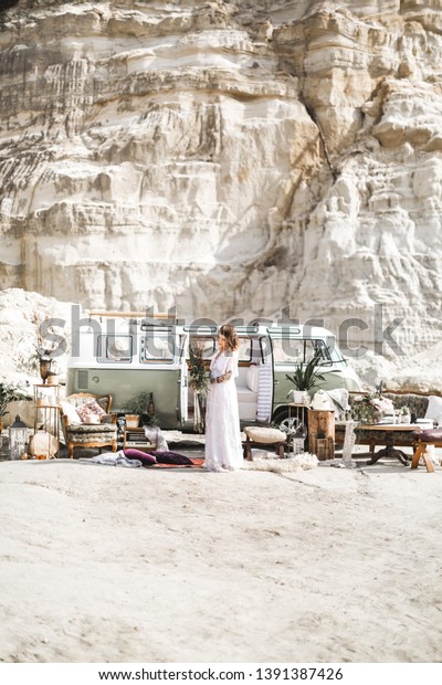 summer, canyon,\
desert, wedding concept - smiling young hippie women in white long\
dress, holding rustic flower bouquet, standing in front of green\
retro minivan car in\
canyon