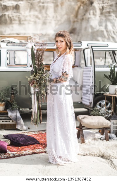 summer, canyon,\
desert, wedding concept - smiling young hippie women in white long\
dress, holding rustic flower bouquet, standing in front of green\
retro minivan car in\
canyon