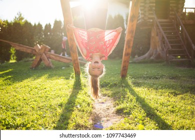 summer children's games and fun outdoors. Happy child girl hanging upside down on wooden Playground on Sunny day - Powered by Shutterstock