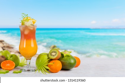 Summer cocktail with various tropical fruits around. Apple, kiwi, tangerine, orange, mango, carambola. Glass of orange drink with blur beach on background - Powered by Shutterstock