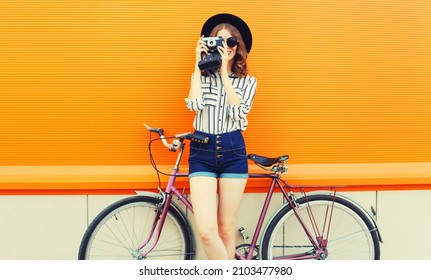 Summer Colorful Portrait Of Happy Smiling Young Woman Photographer Taking A Picture By Film Camera With Bicycle In The City On Orange Background