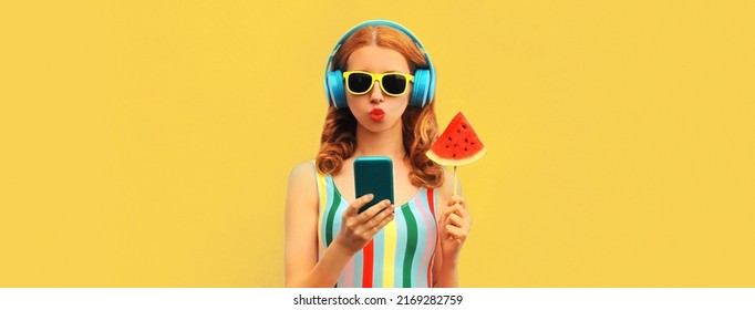 Summer colorful portrait of stylish young woman in headphones listening to music on smartphone with juicy lollipop or ice cream shaped slice of watermelon on blue background - Shutterstock ID 2169282759
