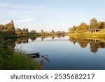 Summer country landscape at sunset. Calm river, reflection of trees in water. Narrow suspension bridge over the Olonka River in the village of Verkhovye, Olonetsky district, Karelia