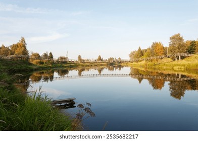 Summer country landscape at sunset. Calm river, reflection of trees in water. Narrow suspension bridge over the Olonka River in the village of Verkhovye, Olonetsky district, Karelia - Powered by Shutterstock