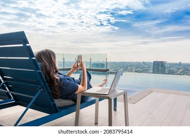 Summer day lifestyle asian woman relax and chill near luxury swimming pool. Asian woman using app on a smartphone and working on a laptop and Blurred background skyscraper, Beautiful sky and clouds.