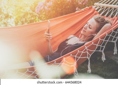 Summer day, young woman lying in orange hammock in park and listening to music on smartphone.Girl blogging, chatting, shopping online, using digital gadget.Lifestyle. Instagram filter. Social network. - Powered by Shutterstock