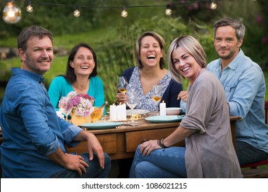 A Summer Evening Of Friends In Their 40s Gather Around A Table In The Garden To Share A Meal And Have Fun Together.